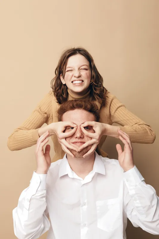 Happy young woman in beige sweater playfully covers eyes of smiling man in white shirt. Studio portrait on beige background. Concept of friendship, fun, joy, and casual lifestyle.