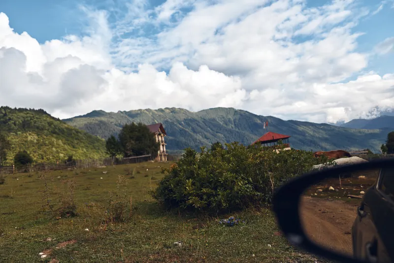 Scenic landscape of rolling hills and countryside seen through a car side mirror, a distant small house, vast blue sky, and open fields conveying travel and exploration.