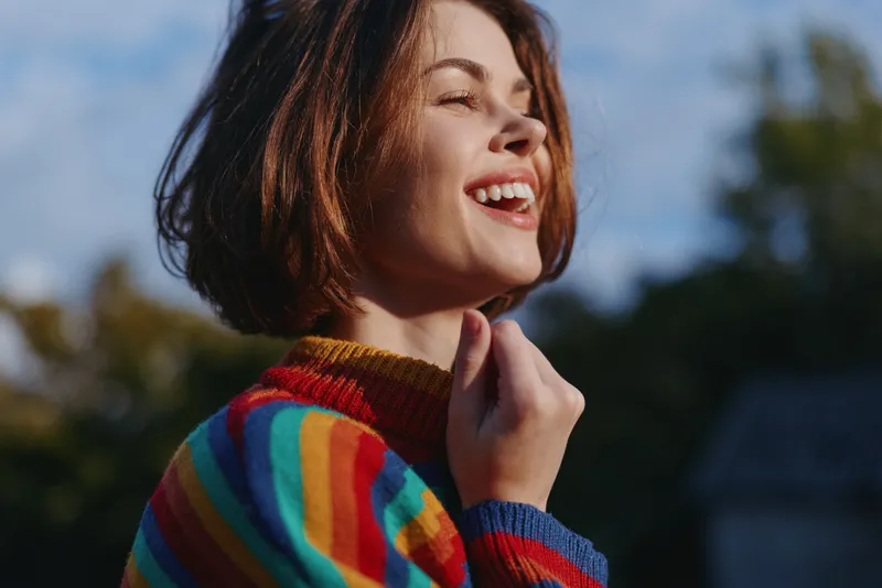 woman smiling in her mid twenties with short brown hair, wearing a colorful striped sweater outdoors in warm sunlight, joyful expression, casual outfit, lifestyle portrait for travel and fashion