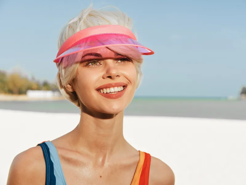 Bright portrait of a smiling woman at the beach wearing a pink visor, sunlit skin, and colorful swimsuit straps, capturing warmth, confidence, and carefree summer glow.
