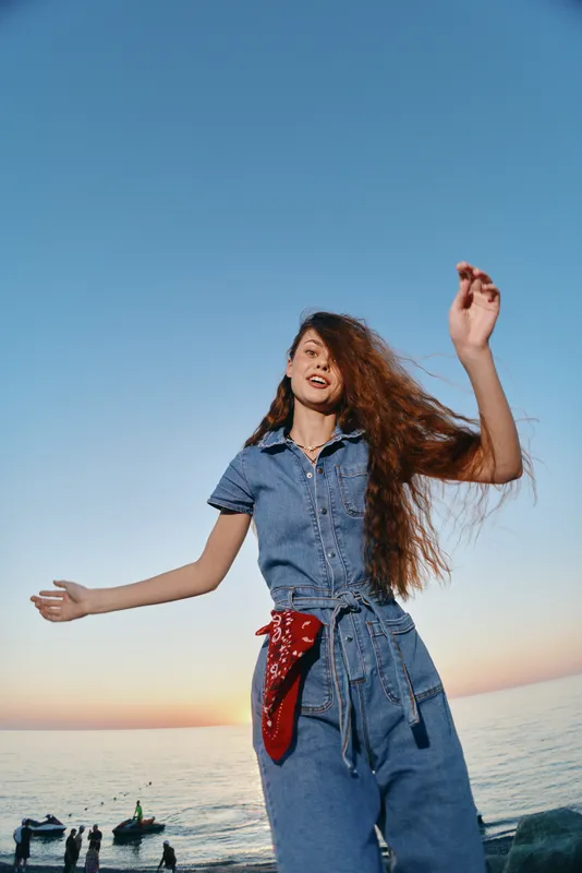 young woman dancing outdoors in denim outfit with long curly hair during sunset by sea with clear sky and boats in background