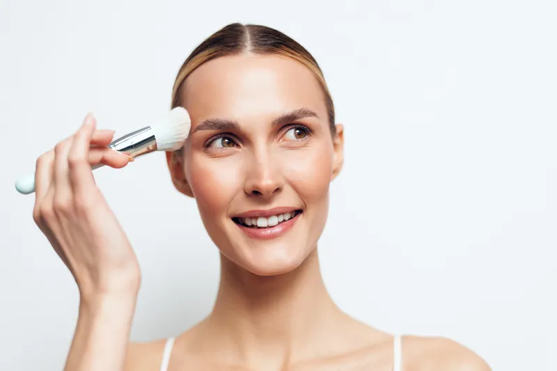 Smiling young woman applying makeup with a brush on her face. Close-up on a white background. Concept of beauty, skincare, and daily routine with natural lighting.