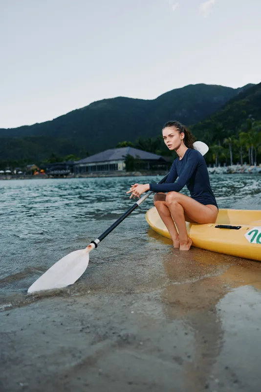 Woman paddling kayak near shore with mountains in background on calm water, wearing navy blue swimsuit, outdoor adventure, summer sport activity.