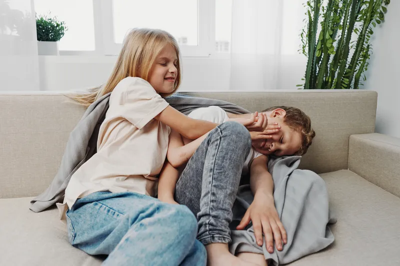 Playful children sitting on a sofa in a bright living room, a girl gently covering a boy's face with her hand. Concept of sibling interaction, family bonding, and childhood moments.