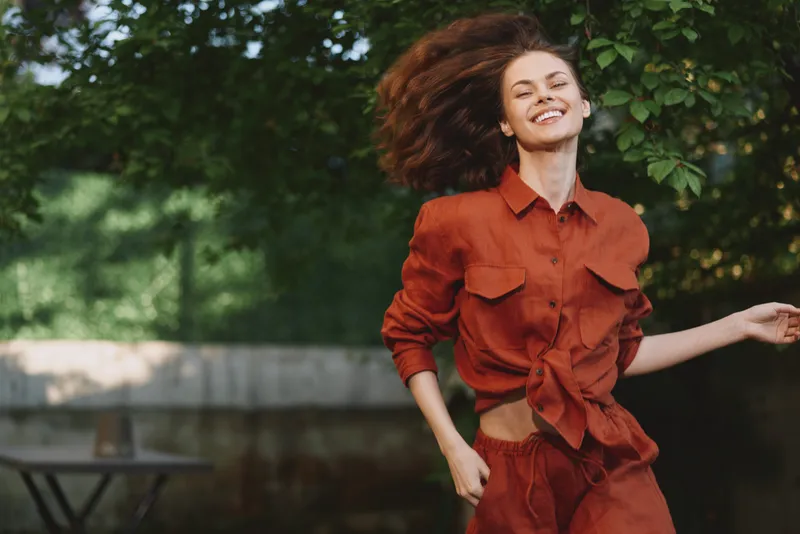 Young woman in a rust shirt and shorts runs joyfully outdoors, hair flowing, smiling, capturing a lively lifestyle moment in a sunny park full of trees and warmth