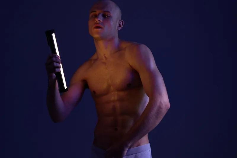 Athletic man posing with a neon light in a dark space, showcasing a strong physique. The composition highlights his confident expression and the interplay of shadows.