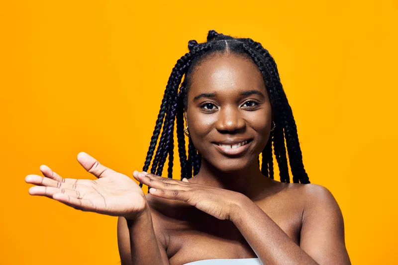 Young Black woman with braided hair smiling against a vibrant yellow background, showcasing joy and confidence in a stylish, casual outfit