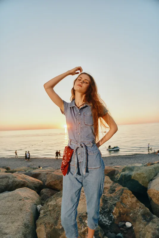 young woman standing on rocky beach at sunset wearing casual denim outfit with relaxed confident posture and calm sea background