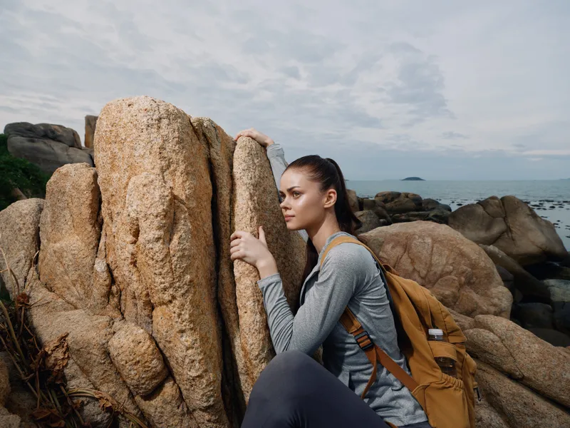 Young woman in outdoor adventure gear resting against a rock by the sea, showcasing determination and exploration