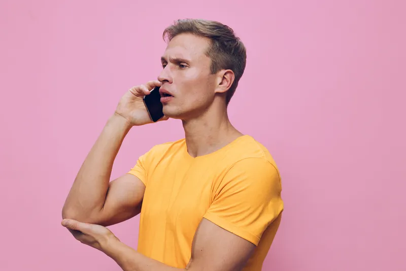 Man talking on mobile phone, surprised expressions, casual yellow t-shirt, short brown hair, clean background, close-up shot, modern lifestyle concept, studio portrait image, colorful, vibrant, indoor