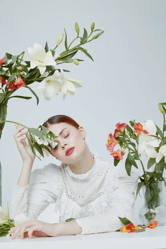 A serene young woman in white attire gently holds a leaf while resting her head on her hand amidst vibrant flowers, conveying tranquility and natural beauty