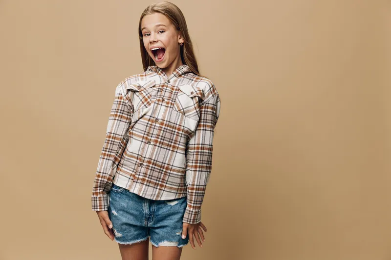 Girl with long hair wearing plaid shirt and denim shorts posing excitedly with open mouth and hands by sides on beige background in studio. Youthful casual fashion concept.