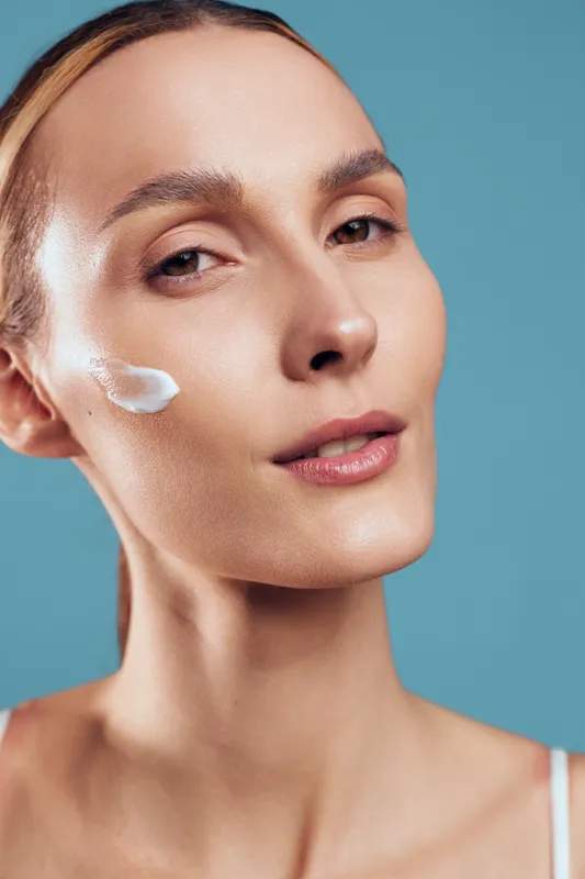 Closeup portrait of woman applying cream on cheek, smooth skin texture, natural makeup, serene expression, blue background, skincare routine, healthy complexion.
