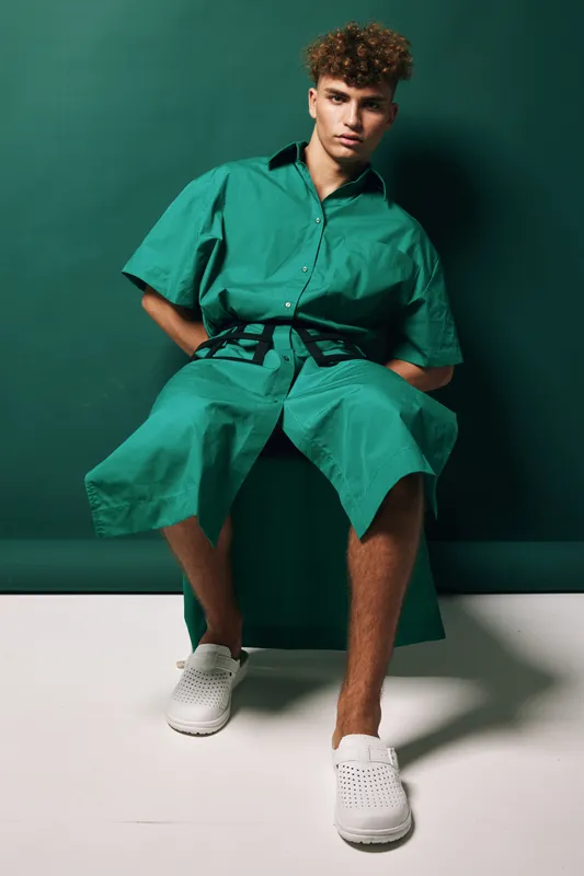 Young man with curly hair wearing green oversized shirt and matching shorts sitting on chair against green wall. Casual fashion, relaxed pose and modern style with white slip-on shoes.