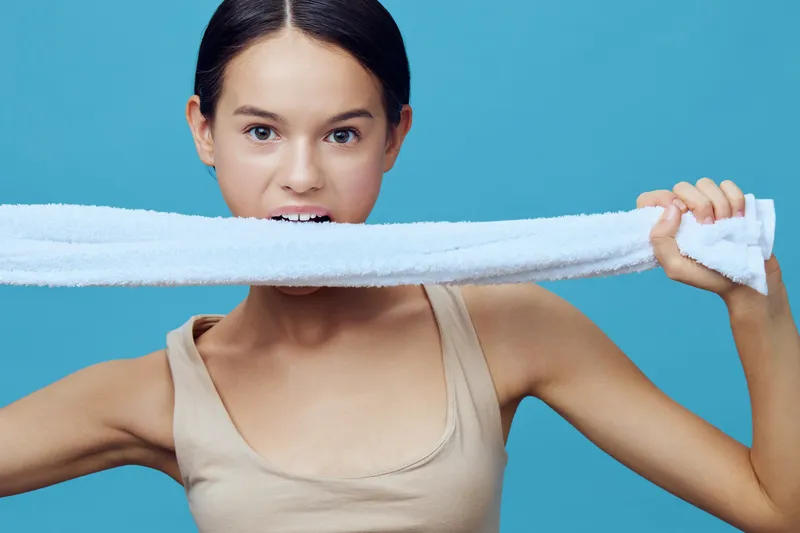 Young girl with brown hair playfully biting a towel against a bright blue background, showcasing a cheerful and energetic attitude, perfect for fitness and wellness themes