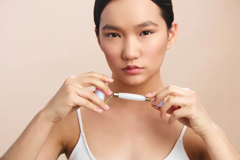 asian woman uses a beauty device for skincare in a clean studio setting. She holds the tool with calm focus, conveying modern selfcare and a thoughtful grooming routine.