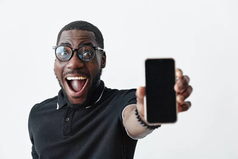 African American man with dark skin and glasses showing smartphone with blank screen, smiling excitedly in casual black shirt on white background for technology and communication.
