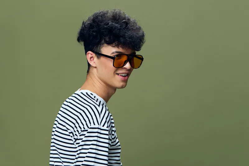 Young man with curly hair wearing sunglasses and striped shirt stands against green background, showing casual style and relaxed expression, portrait in studio lighting.