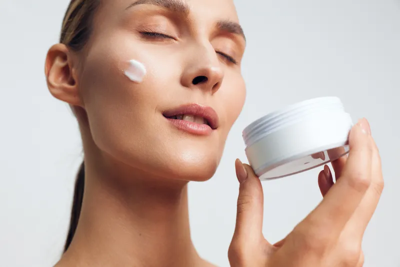 A young woman with clear skin applies moisturizing cream on her cheek while holding an open white jar. The bright background emphasizes skincare and beauty routine concepts.