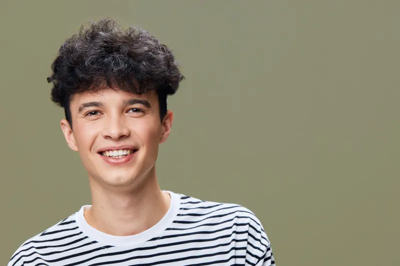 smiling young man with curly hair wearing white and black striped shirt against plain olive background, casual style, positive expression, studio portrait