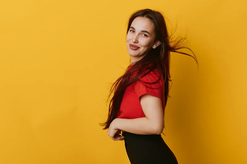 Young woman with long hair wearing red shirt and black pants posing confidently against yellow background with natural smile and relaxed posture for casual portrait.
