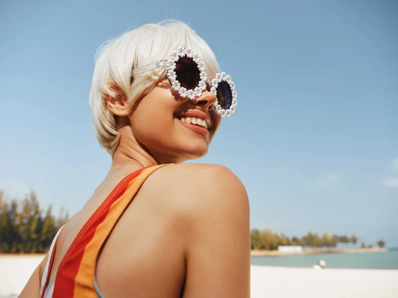 A bright sunlit portrait of a smiling person with short blonde hair, wearing oversized pearl sunglasses, captured on a clear beach day with blue sky.