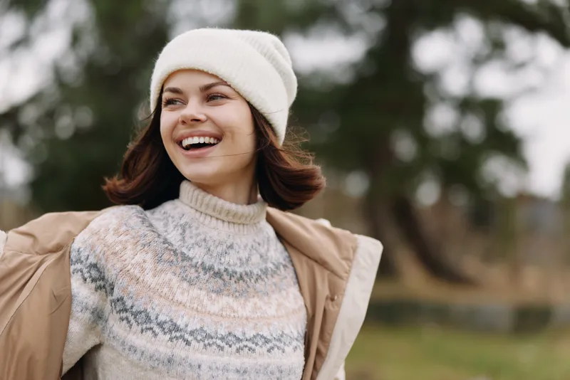Woman outdoors in a cozy sweater and beanie, smiling warmly, with a light jacket in a park setting during autumn with a gentle breeze