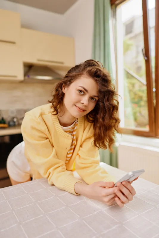 Woman at home leaning on a table with a phone, casual cozy kitchen scene, warm yellow sweater, relaxed lifestyle portrait, natural light and warm mood.