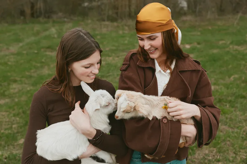 Lifestyle scene with two women outdoors, holding fluffy goats, a gentle moment of bonding and care, warm clothing, grassy field, daytime light, authentic rural companionship and joy