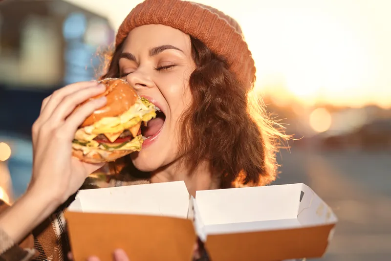 Woman eating burger from streetfood box outdoor, candid joyful portrait capturing authenticity and golden hour glow, candid lifestyle moment with mindful living and emotional storytelling.