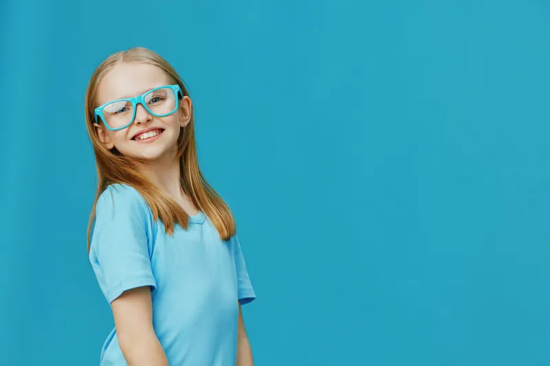 Smiling girl with glasses in a blue shirt against a bright blue background, exuding happiness and confidence, perfect for children s fashion and educational themes