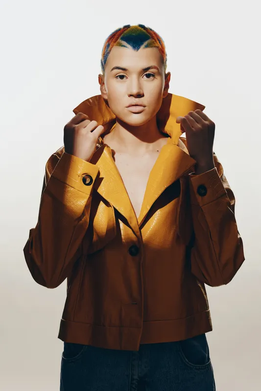 young woman with colorful hair wearing a yellow jacket stands confidently against a light background, showcasing modern style and bold fashion choices