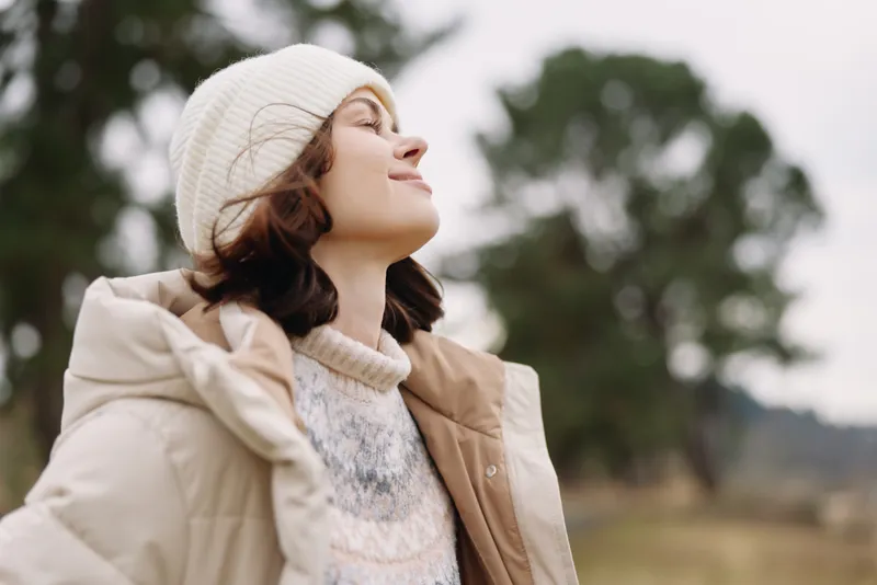 Woman in a warm coat and beanie outdoors, smiling and looking up, with a soft natural background and autumn winter fashion mood.