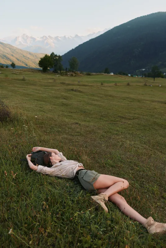 Woman lifestyle outdoors lying on grass field with arms raised relaxed pose casual clothing natural light mountain background summer day