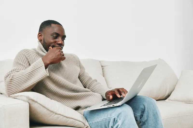 Thoughtful African man in a cozy sweater working on a laptop at home, smiling softly, showcasing productivity and comfort in a neutral toned living room