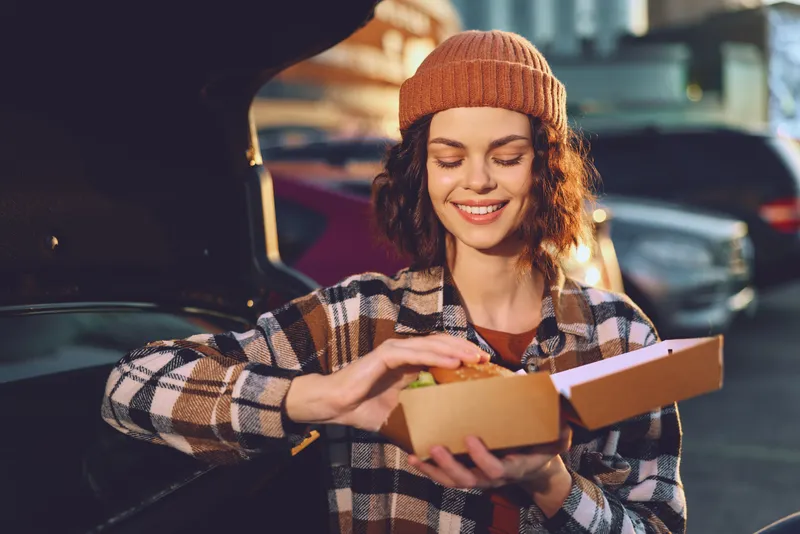 Woman with food takeaway at car trunk, smile in beanie and plaid jacket during evening golden hour glow, candid lifestyle portrait with authenticity, mindful living vibe.
