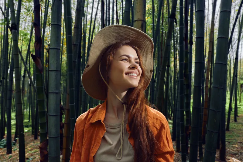 Woman bamboo forest hat smiling in orange shirt, portrait in nature outdoors, traveler enjoying sunlight and peaceful green environment during eco hiking escape in serene woodland.
