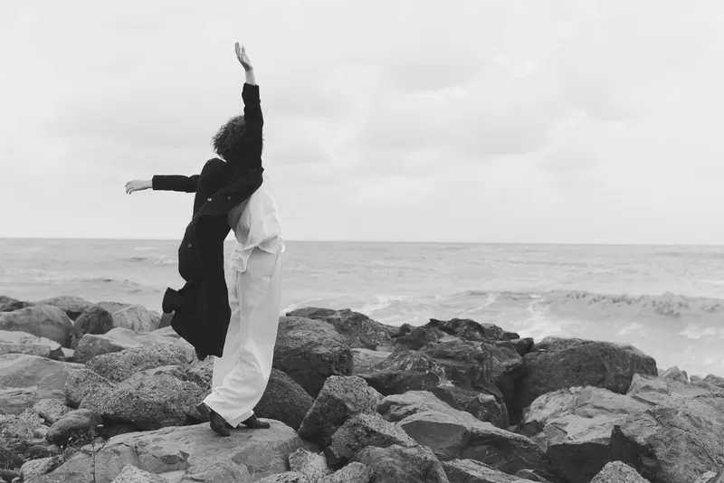 Dance, couple on shore with waves, embrace and lift arms on rocky beach, monochrome scene capturing movement, joy and freedom by the sea