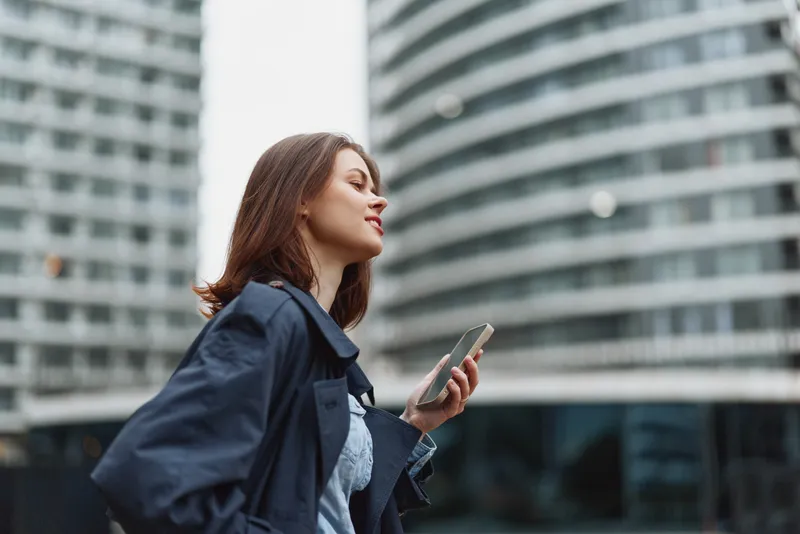 A confident woman in a navy jacket walks outdoors, holding a smartphone, looking ahead with a calm smile. A modern cityscape provides a dynamic, urban backdrop.