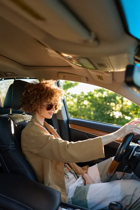 A driving woman sits in a retro car with sunglasses, relaxed posture, and curly hair. Daylight streams through the interior, creating a fashion forward lifestyle scene of a stylish road trip.