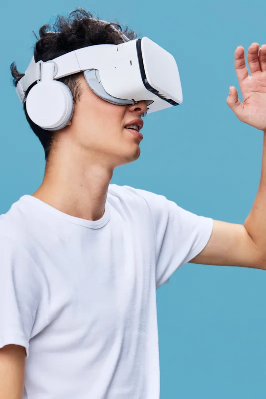 Young man wearing white t-shirt and VR headset with headphones exploring virtual reality, interacting with digital environment on a blue background.