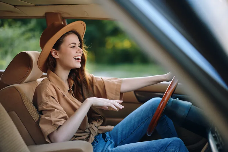 Smiling young woman driving a vintage car, wearing a hat and casual clothing, exuding joy and freedom in a natural green background.