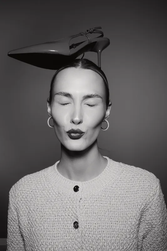 Elegant white woman with pale skin and closed eyes balances a shiny high heel shoe on her head. Black and white portrait with calm expression and minimalist fashion concept.
