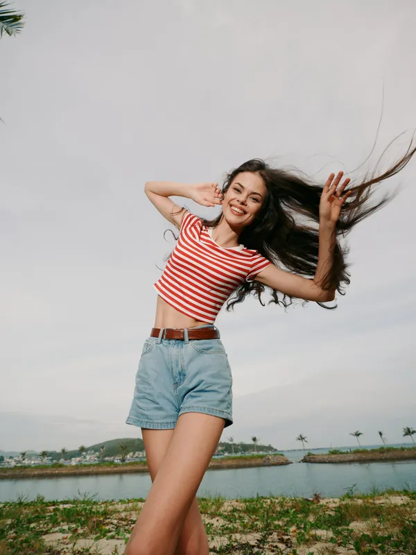 A joyful young woman dances on a seaside trail, wearing a striped crop top and denim shorts, hair flowing freely as she smiles beneath a bright, clear sky.
