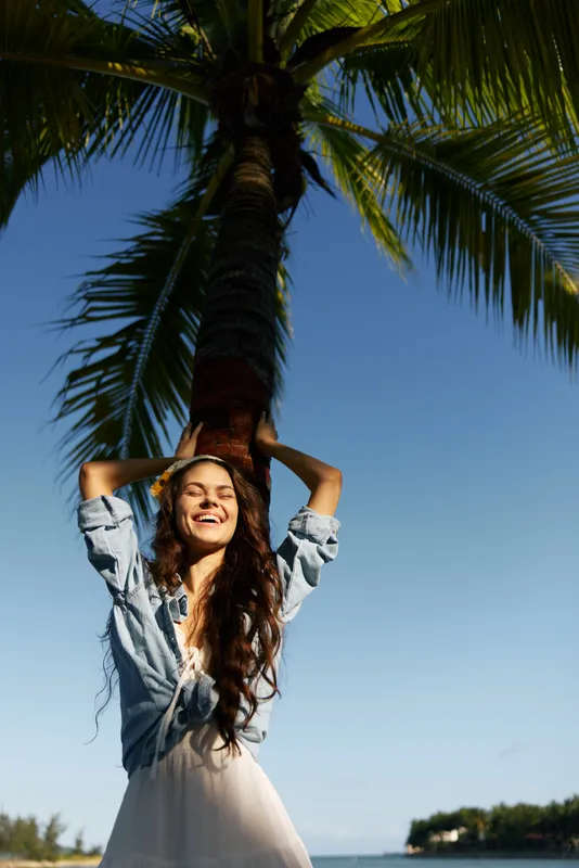 smiling woman standing under palm tree wearing summer dress and shirt, enjoying sunny day by the sea with clear blue sky and tropical background