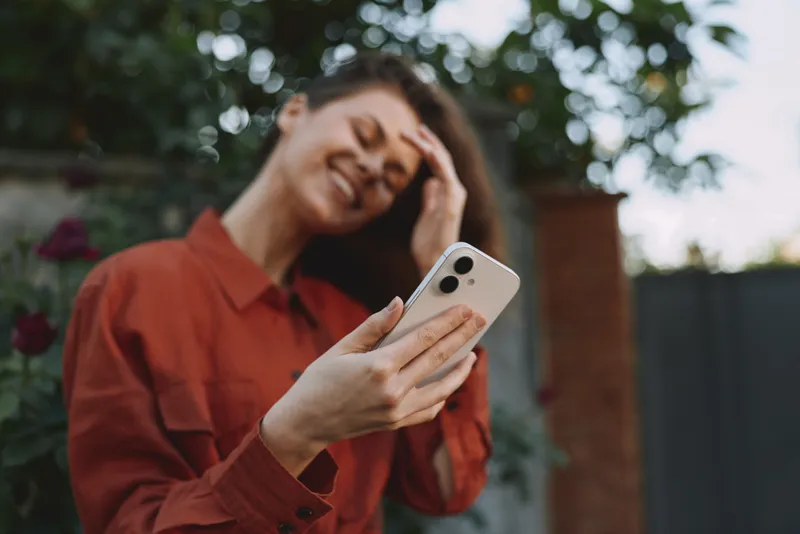 A woman enjoying outdoor lifestyle, taking a selfie in a garden setting, wearing a red shirt, smiling, capturing a bright candid moment in natural light
