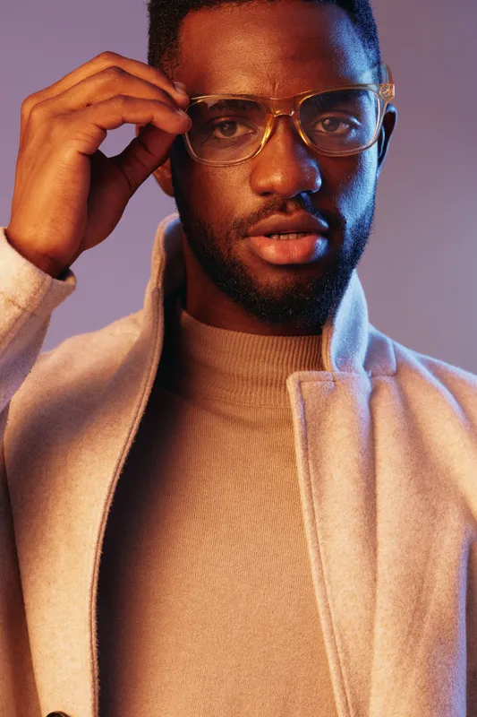 Stylish young Black man in glasses wearing a beige coat against a warm gradient background, exuding confidence with a thoughtful expression