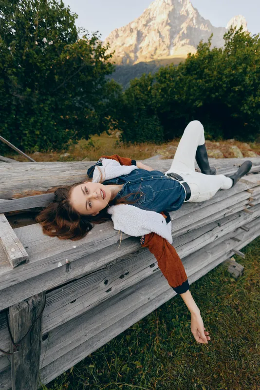 Woman outdoors on wooden bench by mountain, relaxed and smiling in casual denim jacket and white pants. Nature portrait with rustic scenery, meadow and peaceful mood.