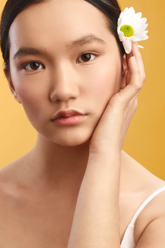Asian woman with a flower in her hair stands against a warm yellow background, presenting a calm, elegant portrait. Soft lighting highlights smooth skin and a thoughtful expression, creating a serene
