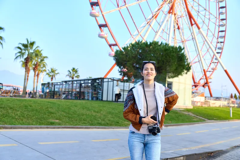 Woman with camera strolling near a ferris wheel at a seaside park, casual jacket and jeans, capturing vibrant travel moments in a lively outdoor setting